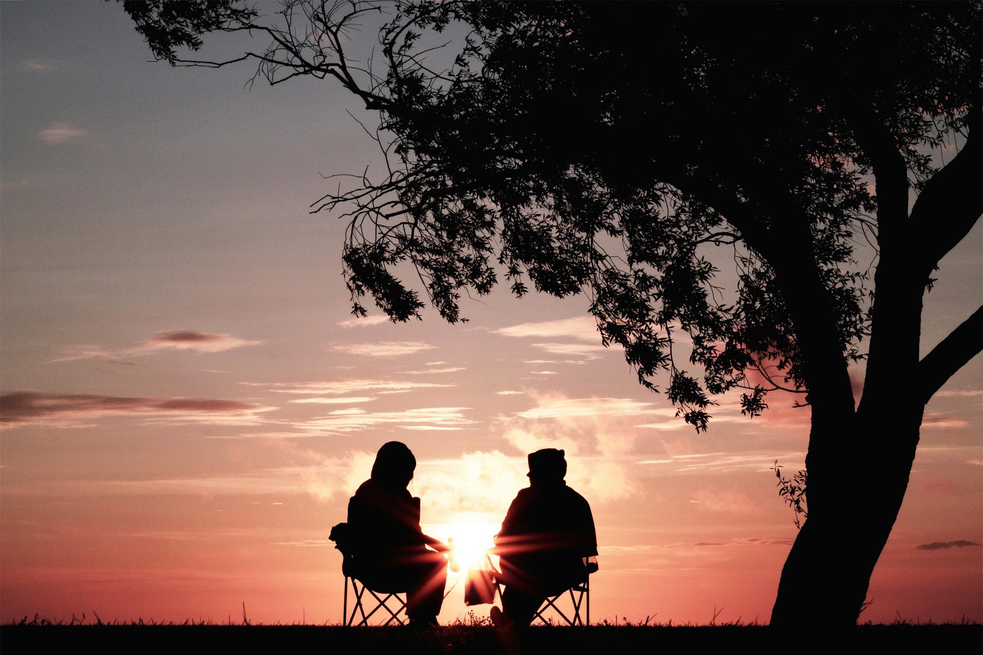 A silhouetted couple sitting on outdoor chairs looking into the sunset
