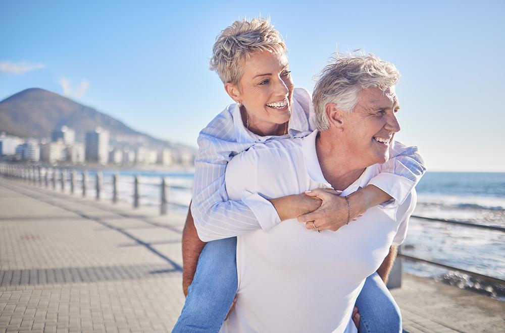 a happy elderly man piggybacking an elderly woman on a beach